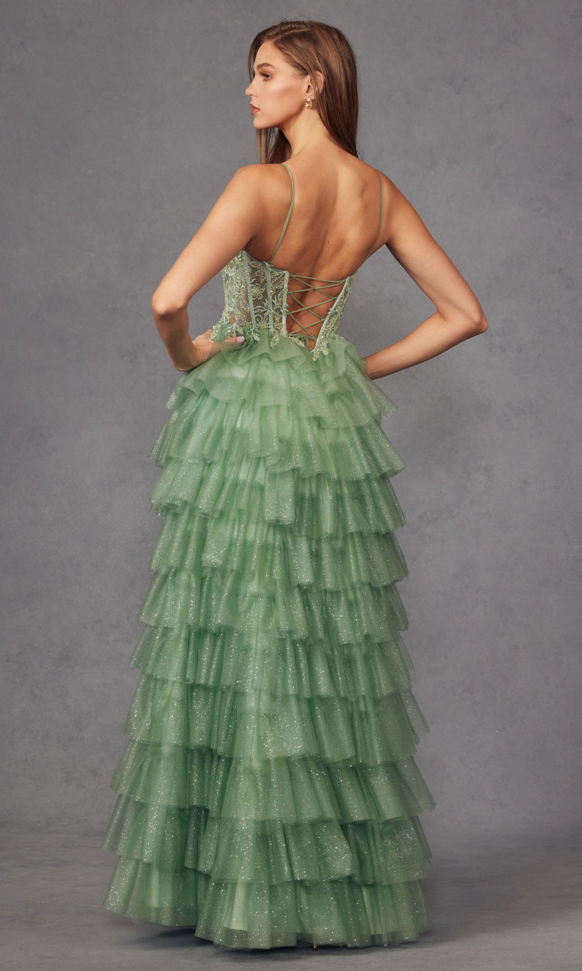 A woman in the Juliet JT2458A Ruffled Glitter Tulle Prom Dress with a light green, tiered skirt and lace-up corset back stands facing away against a plain gray background.